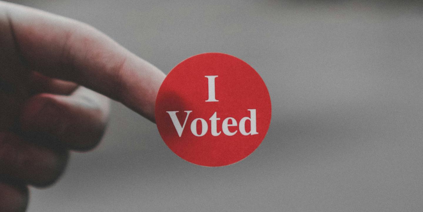 Finger holding up a red "I voted" sticker
