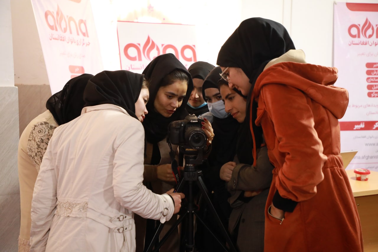 A group of women in hijabs huddle around a single camera on a tripod.