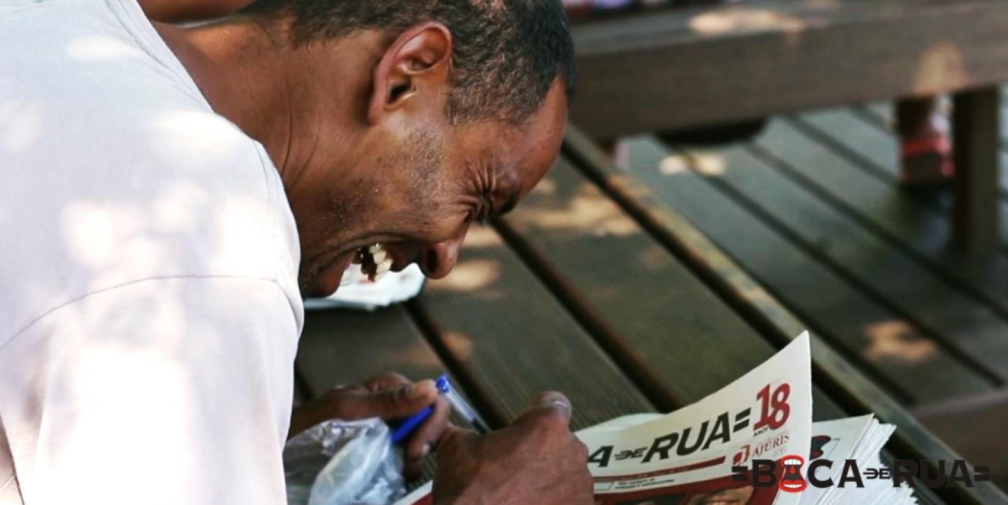 Homem sorrindo segura pilha de exemplares do jornal Boca de Rua