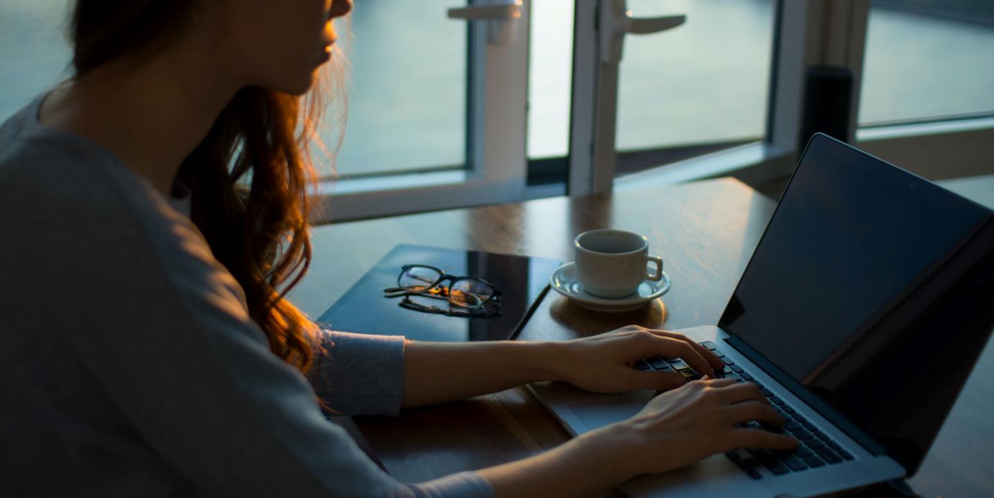 Una mujer trabaja en su computadora portátil junto a una taza de café, un cuaderno y vasos.
