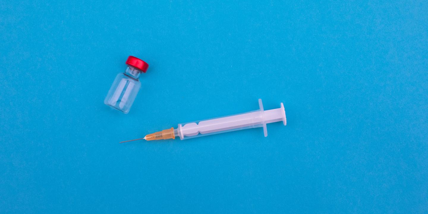 A needle sits next to a small vial against a plain blue background