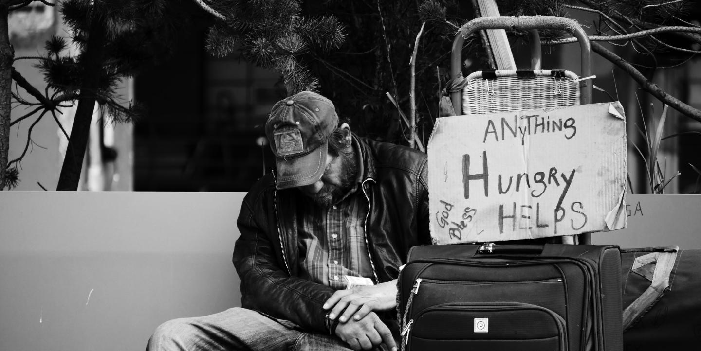 A man sitting outside with his suitcase and a sign that reads: "Hungry, anything helps"