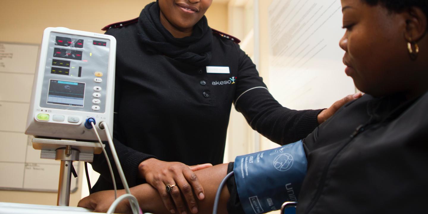 A Black woman smiles, facing the camera, as she takes a sitting Black woman's blood pressure.