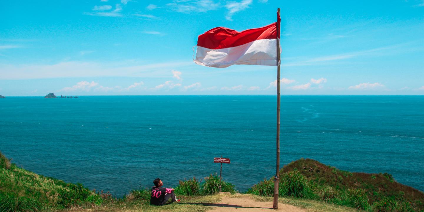 A man sitting at the edge of a cliff looking at a red and white flag. 