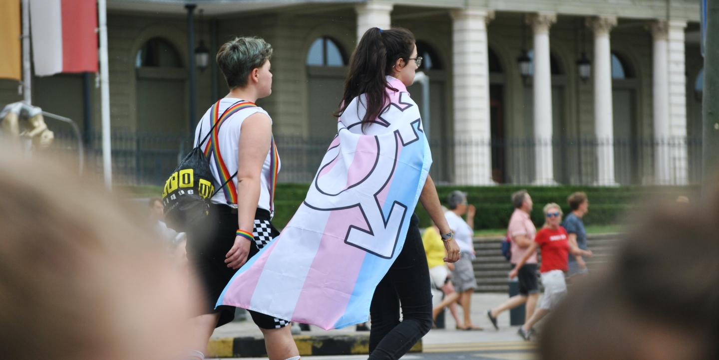 Two people walking and one is carrying a trans flag on their back. 