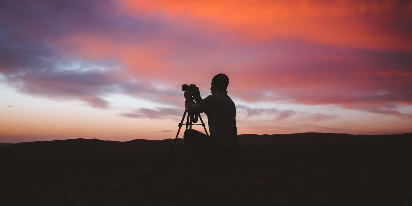 A photographer standing in front of the sunset and taking a picture.