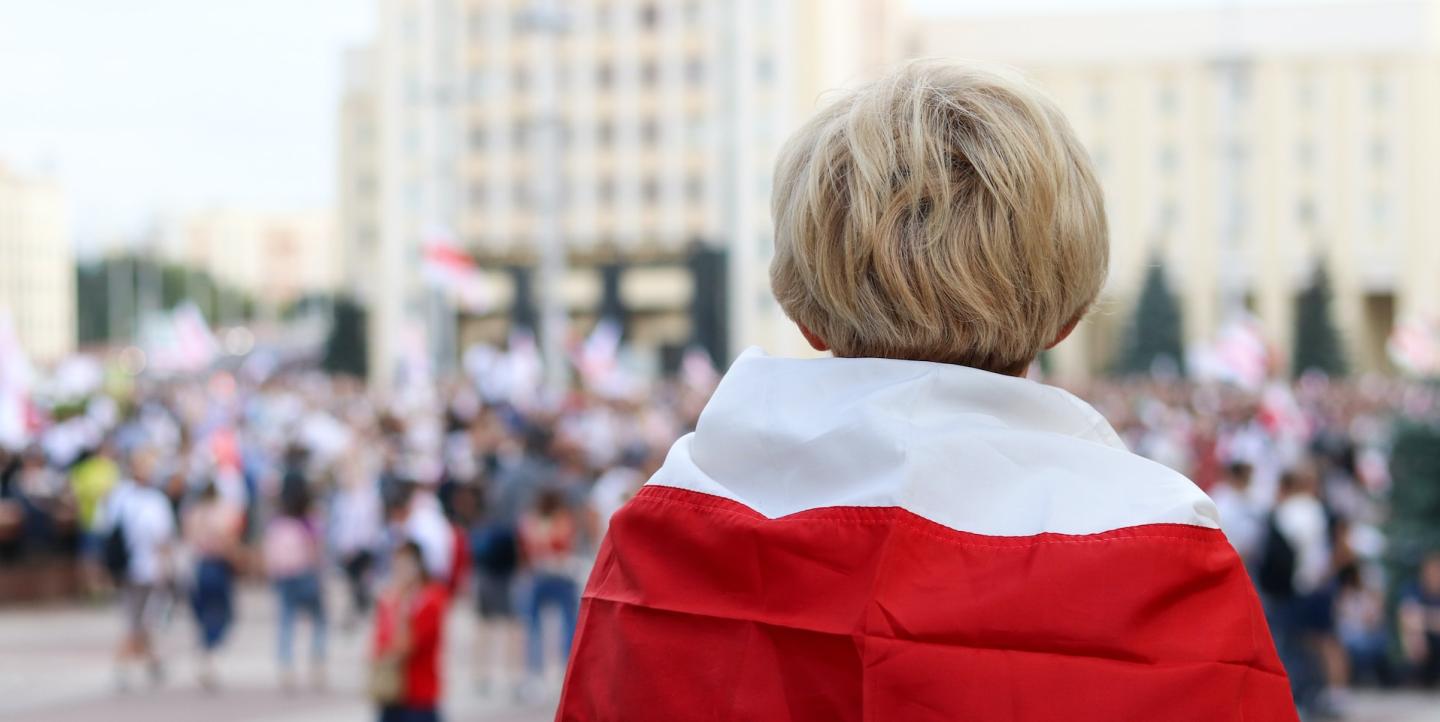 Завернутый в белорусский флаг человек стоит спиной к зрителям на фоне протестов