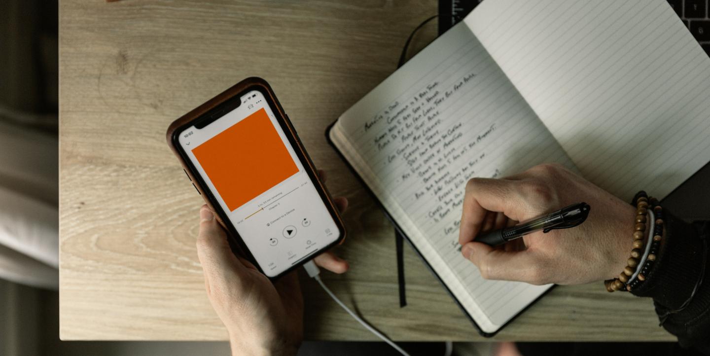 Hombre escribiendo en un cuaderno con un podcast abierto en el teléfono a su lado