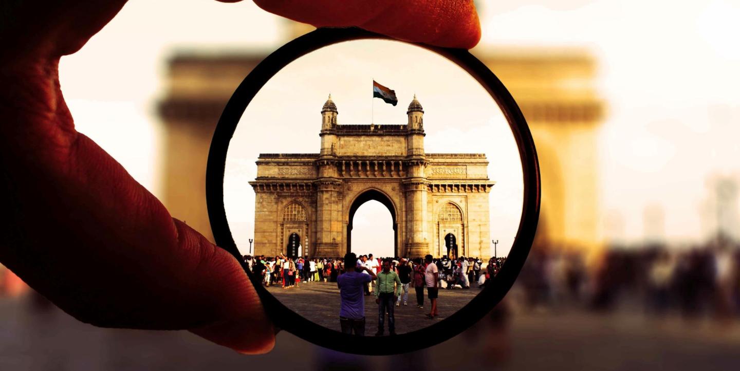 Man holding a magnifying glass in front of an arch with the Indian flag flying above it