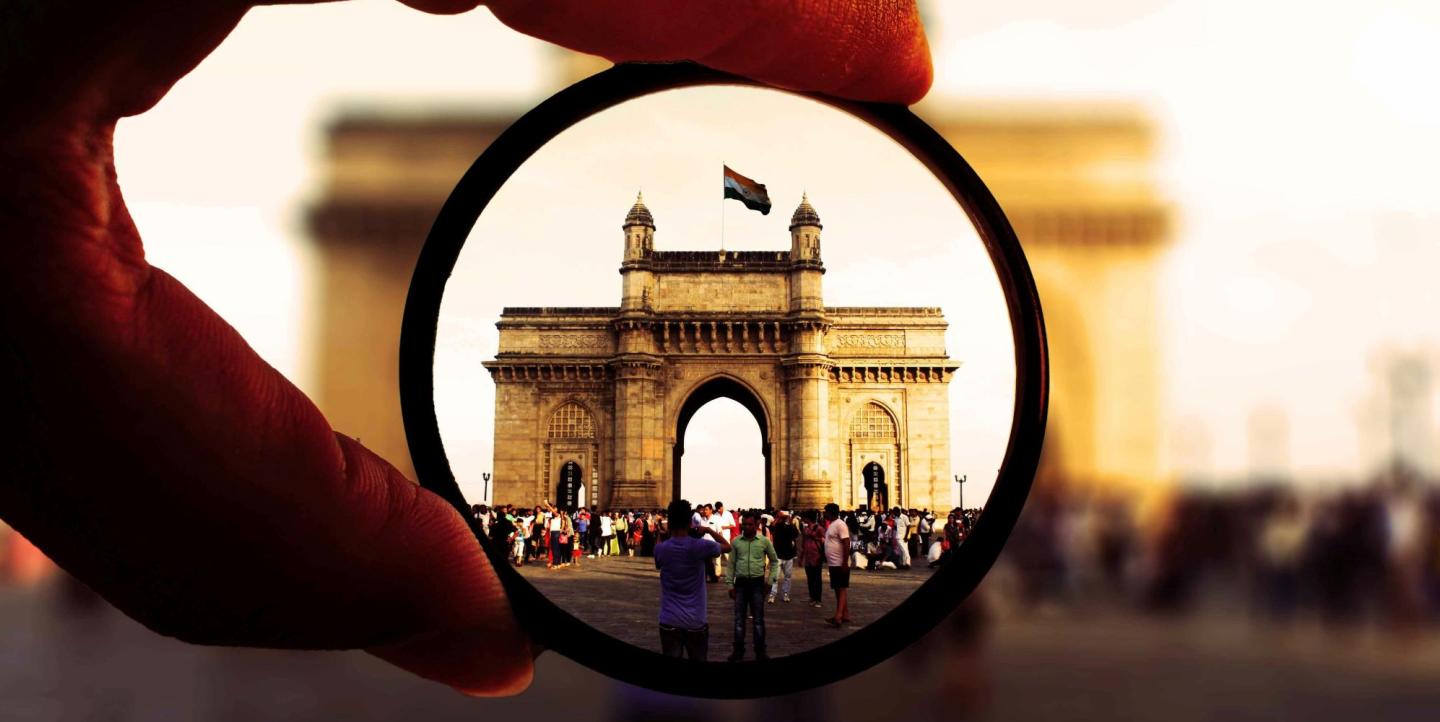 Man holding a magnifying glass in front of an arch with the Indian flag flying above it