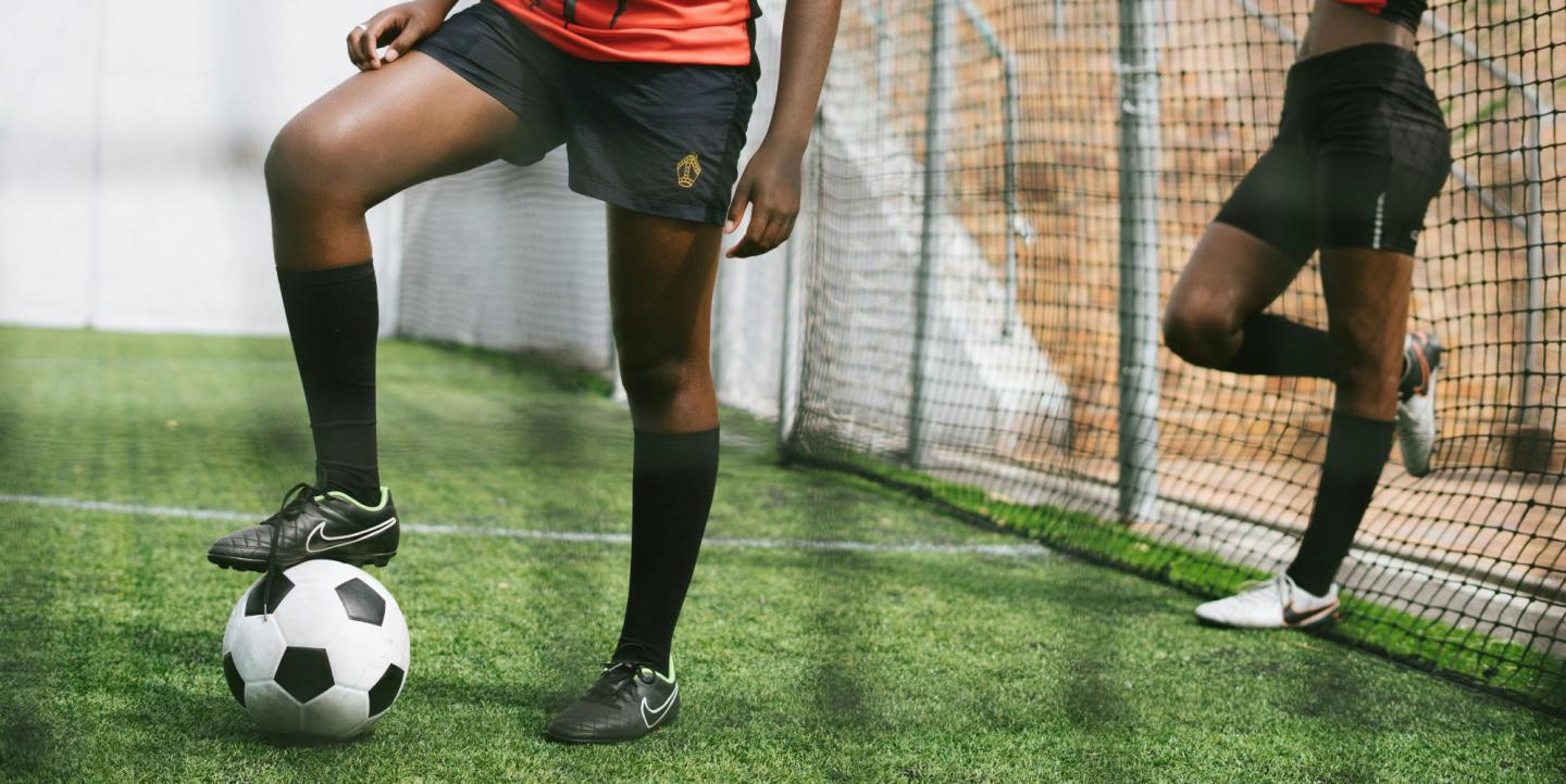 Two women playing soccer, one leaning against the fence and the other with her foot on the ball