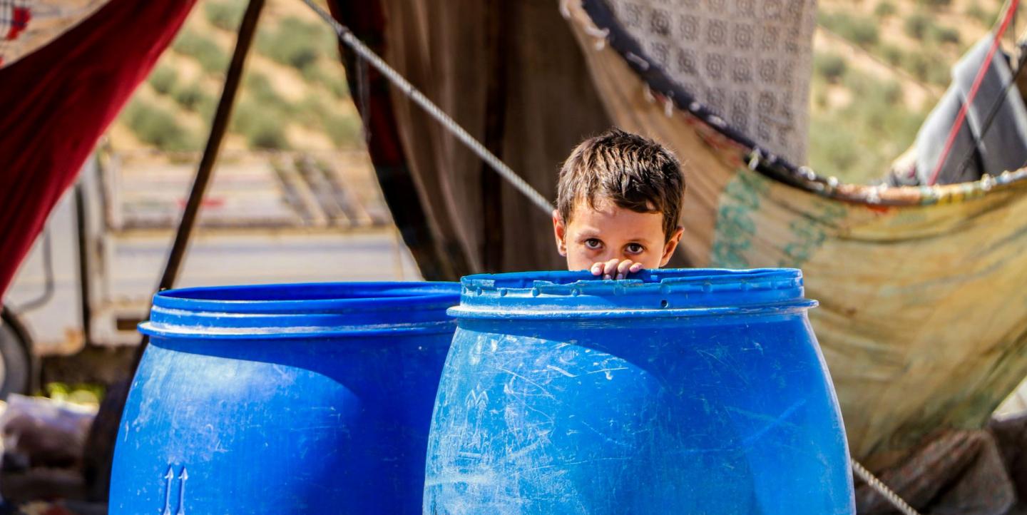 A young boy hiding behind blue barrels in a refugee camp