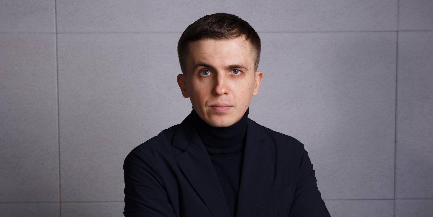 Mykhailo Tkach face caméra portant un col roulé noir et une veste noire devant un fond gris