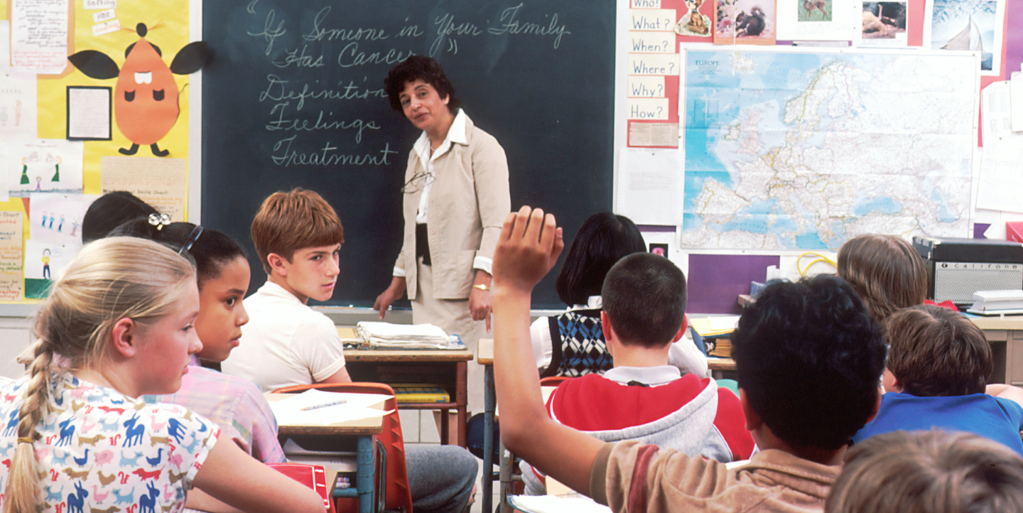 Children in a classroom looking at one student who is raising his hand.