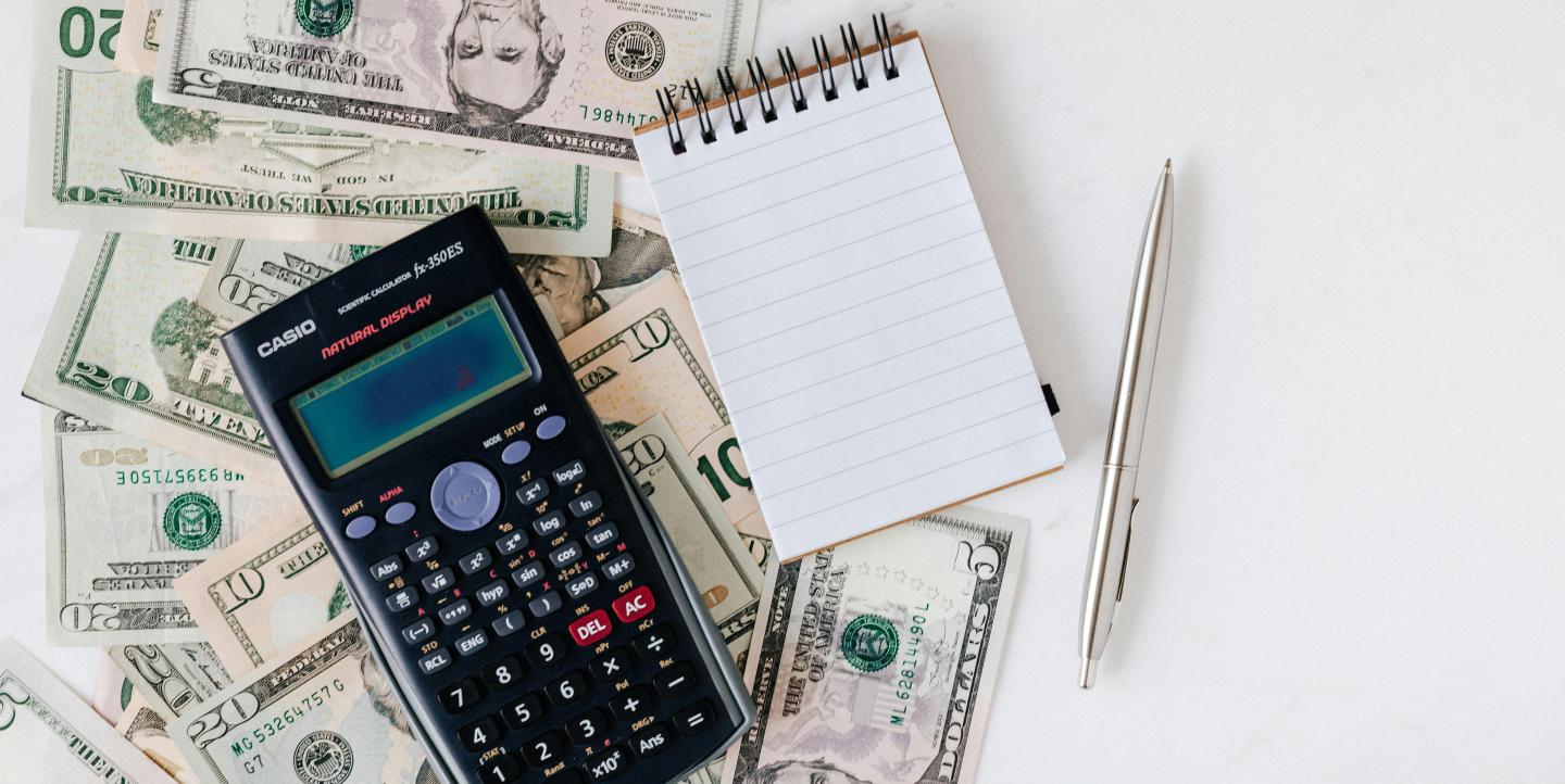 A calculator, pin and notepad on top of scattered cash.
