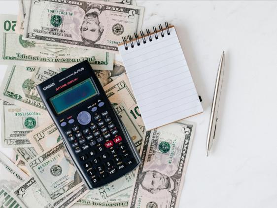 A calculator, pin and notepad on top of scattered cash.