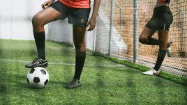Two women playing soccer, one leaning against the fence and the other with her foot on the ball