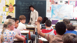 Children in a classroom looking at one student who is raising his hand.