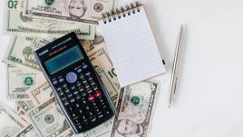 A calculator, pin and notepad on top of scattered cash.